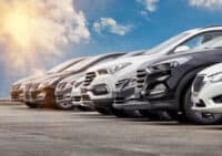 A low-angle shot captures a row of modern SUVs and crossovers, predominantly in white, grey, and black, parked on an asphalt surface under a bright blue sky with scattered clouds. A strong sun glare is visible from the top left, illuminating the scene. The vehicles are arranged in a diagonal line, showcasing their front and side profiles in a clean, optimistic setting.