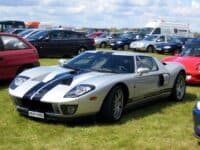 A silver Ford GT with wide black racing stripes on its hood and roof is parked on a green grassy field under a partly cloudy sky. The car is angled towards the left, showcasing its sleek design, front headlights, and side vents. Numerous other cars, including sedans and vans, are visible parked behind it in the background.