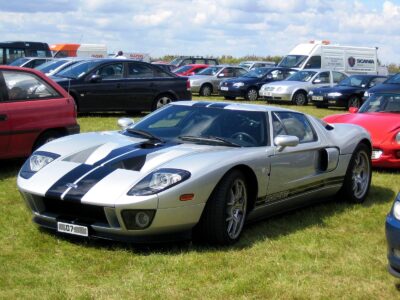 A silver Ford GT with wide black racing stripes on its hood and roof is parked on a green grassy field under a partly cloudy sky. The car is angled towards the left, showcasing its sleek design, front headlights, and side vents. Numerous other cars, including sedans and vans, are visible parked behind it in the background.