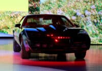 A black Pontiac Trans Am, a replica of the Knight Rider KITT car, is parked on a highly reflective floor. The car's body is glossy, reflecting vibrant blue and red lights from the surrounding environment. The iconic red scanner light across the front grille is illuminated, glowing brightly. The background features abstract, blurred patterns of green, yellow, and blue light, suggesting a display or stage setting.