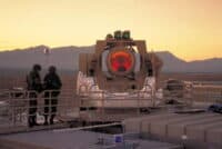 Two soldiers in military camouflage stand on a platform observing a large, light brown laser weapon system. The weapon system has a prominent, red glowing emitter and various sensors on top. The scene is set in a vast, arid desert landscape with distant mountains, under a warm, golden sunset sky. The soldiers appear to be looking at a device.