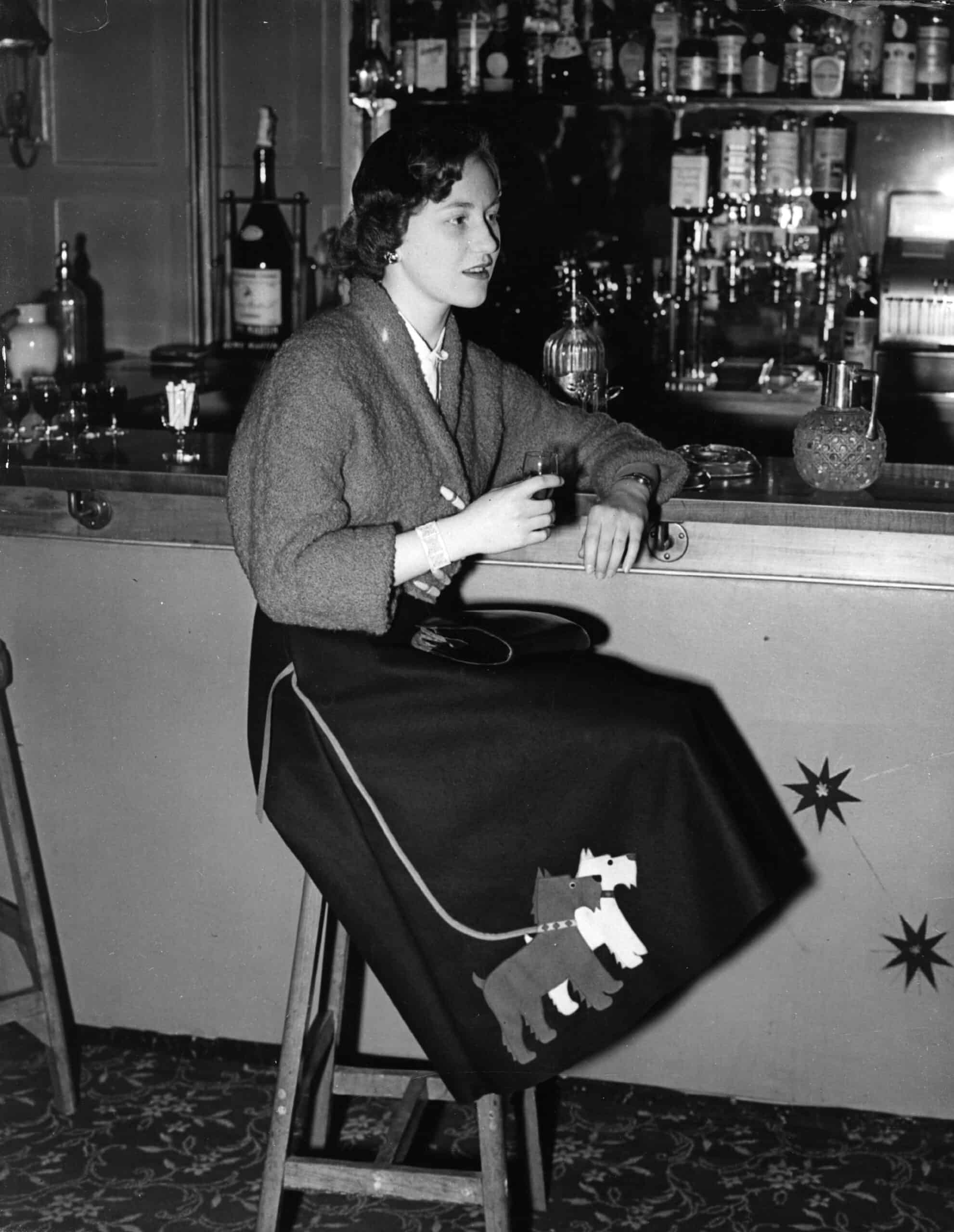 A young woman sits at a bar wearing a poddole skirt and sweater. 