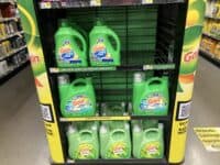 A green and yellow retail display stand is filled with large, green plastic bottles of Gain laundry detergent and fabric softener. The top shelves hold Gain +ultra OXI detergent, the middle shelves feature Gain +odor defense detergent, and the bottom shelves display Gain fabric softener. Some shelves show empty slots, and price tags are visible, along with a QR code on the side of the display.