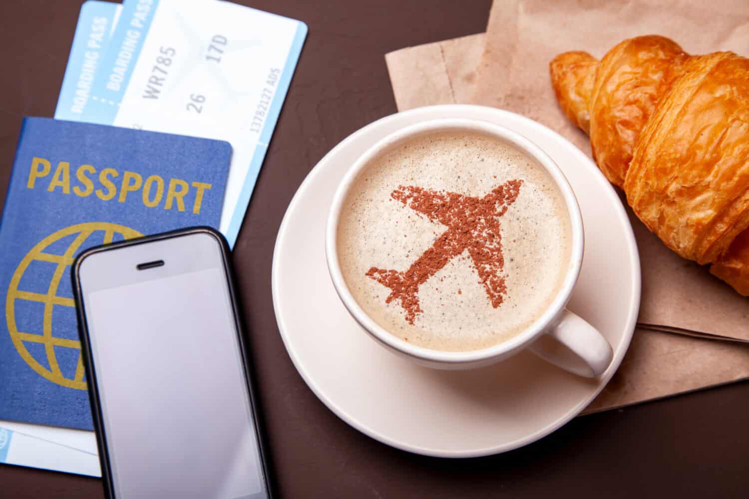 Mug of coffee with an airplane on the foam. Morning coffee with croissant in flight. Paspor and ticket with smrtrfonom and cup of coffee