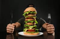 A person, visible from the chest down, sits at a dark table, holding a knife in their left hand and a fork in their right. In front of them, centered on a white plate, is an extremely tall, multi-layered hamburger with sesame seed buns. The burger consists of at least five meat patties, layered with melted cheese, fresh green lettuce, and sliced red tomatoes. The background is dark and out of focus.