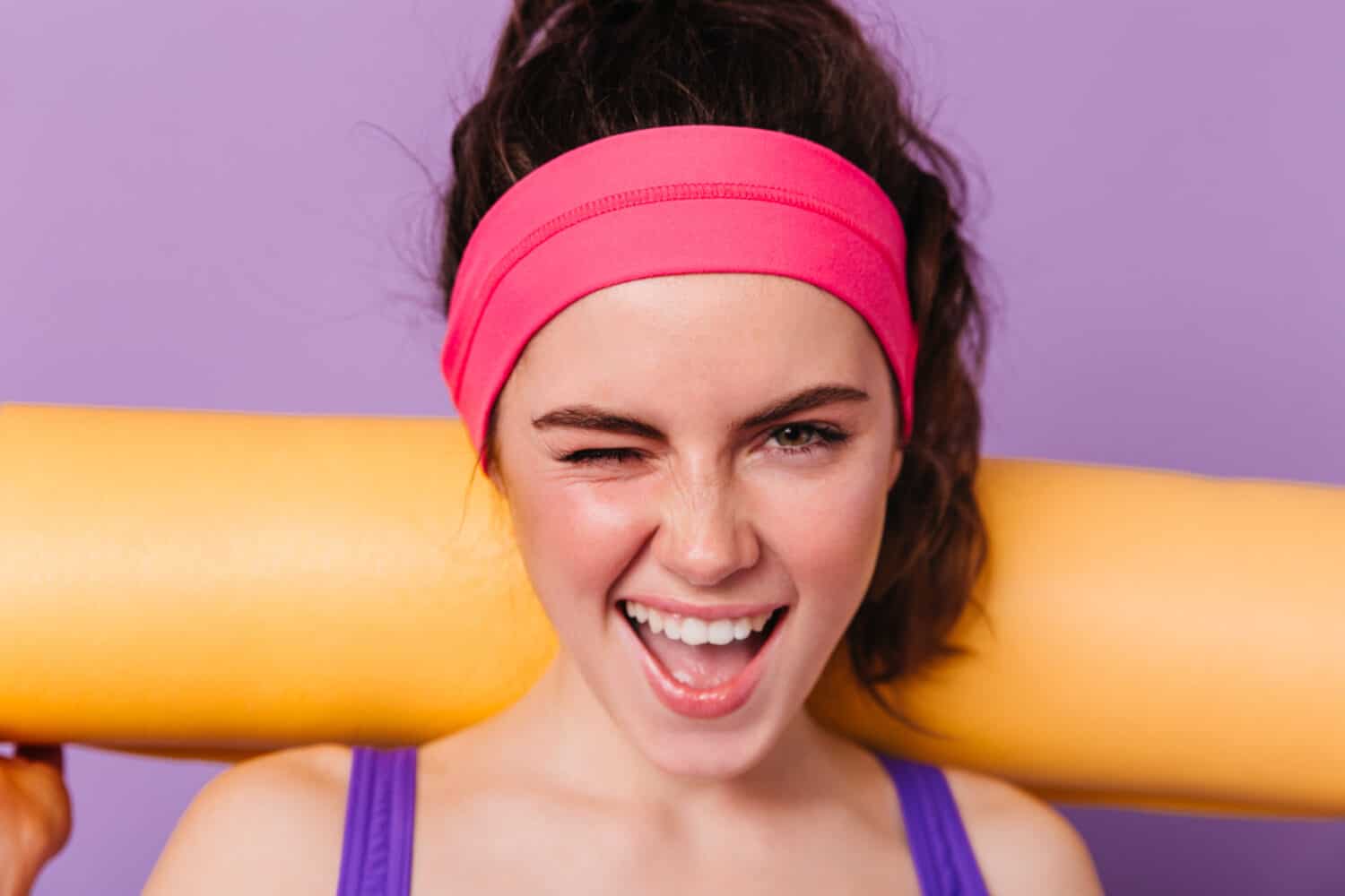 Positive girl athlete in pink headband smiles and winks on purple background with orange mat for fitness