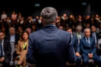 View from behind a man with short, gray hair, wearing a dark blue suit and light blue shirt, speaking to a large, blurred audience. The speaker's back is to the viewer, facing a dimly lit room filled with rows of seated people, some of whom are partially visible in soft focus. The overall composition suggests a professional conference or seminar setting.