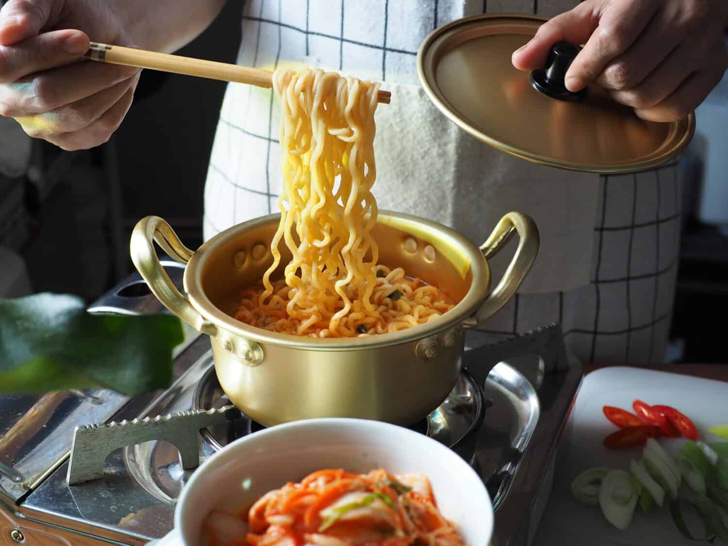 People using chopsticks to cook ramyeon or Korean instant noodles soup in double handle Korean yellow aluminum pot on gas stove over wooden table with bowl of kimchi, slided red chili, scallion.