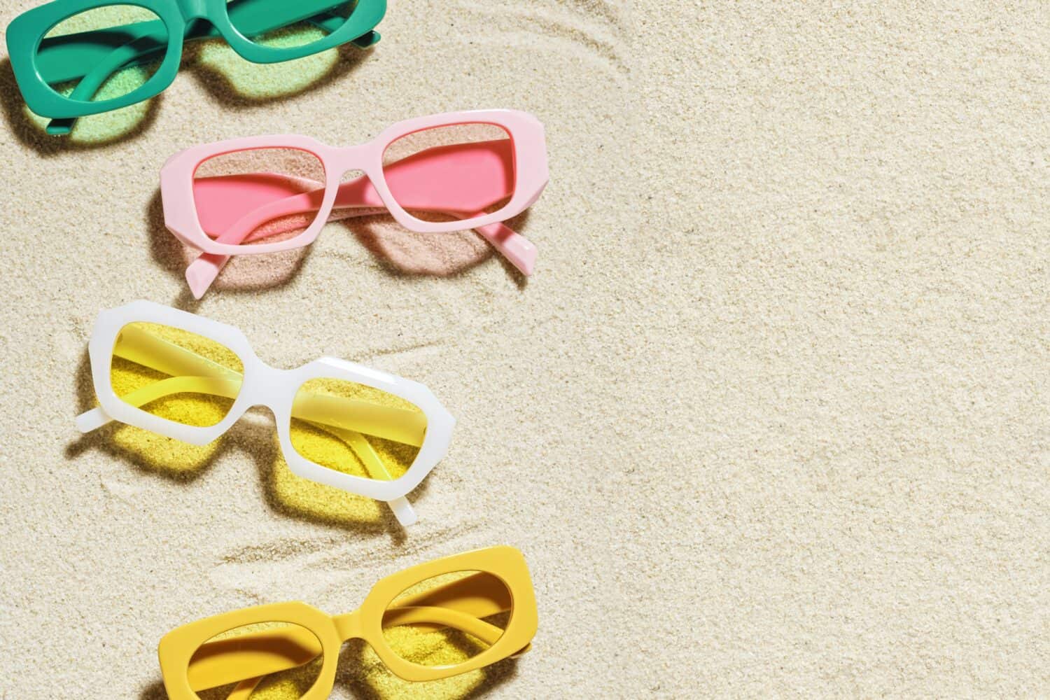 Set of colorful sunglasses on beach sand background at sunlight with shadow. Summer fashion eyeglasses with colored glass. Summer vacation, summer rest concept. Minimal style flat lay, copy space