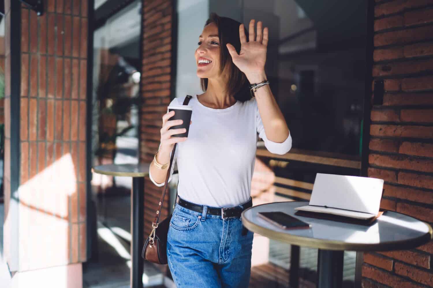 Elegant smiling female in casual clothes with cup of coffee standing at small table outside cafe looking away and waving hand