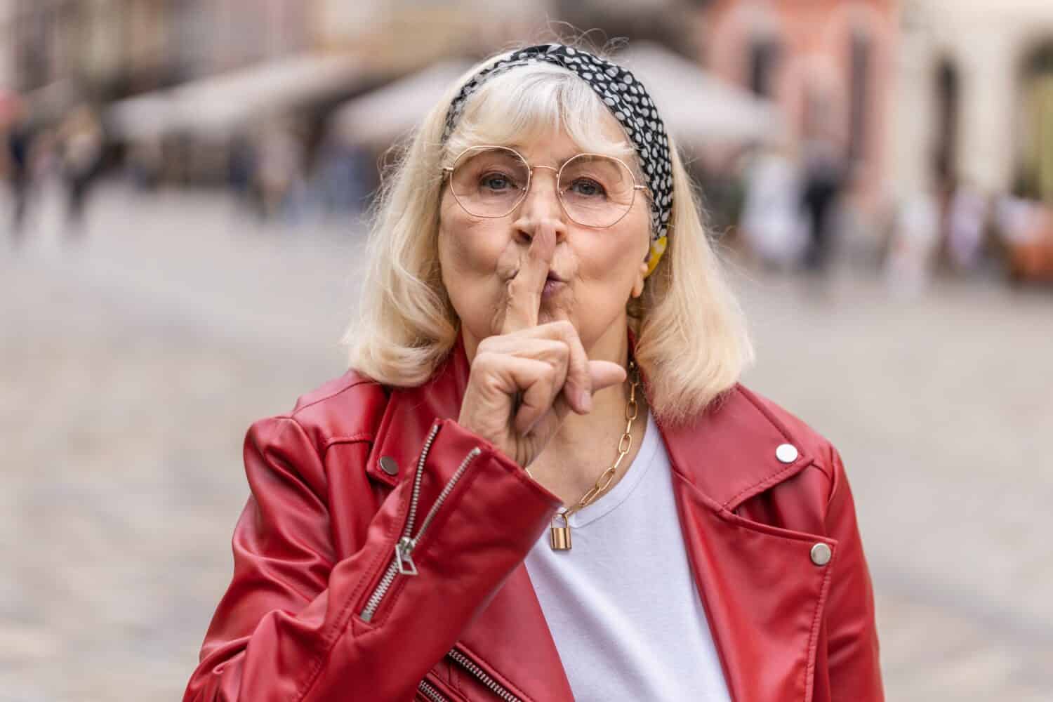 Shh be quiet please. Senior woman presses index finger to lips makes silence hush gesture sign do not tells gossip secretoutdoors. Elderly grandmother walking in urban city street. Town lifestyles