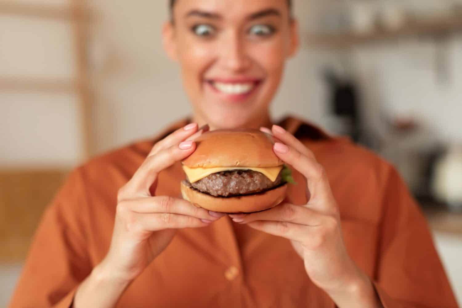 Fast food addiction. Portrait of hungry woman holding burger and looking at it with wide open eyes, enjoying delicious juicy snack, sitting in kitchen, selective focus