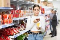 A young woman with dark hair tied back stands in a supermarket aisle, holding a red smartphone in her left hand and a white and yellow product container in her right hand. She is looking intently at her phone. The aisle behind her is filled with shelves displaying various packaged food items, predominantly instant noodle cups and snack bags in red and orange packaging. Two other shoppers are visible in the blurred background on the right.