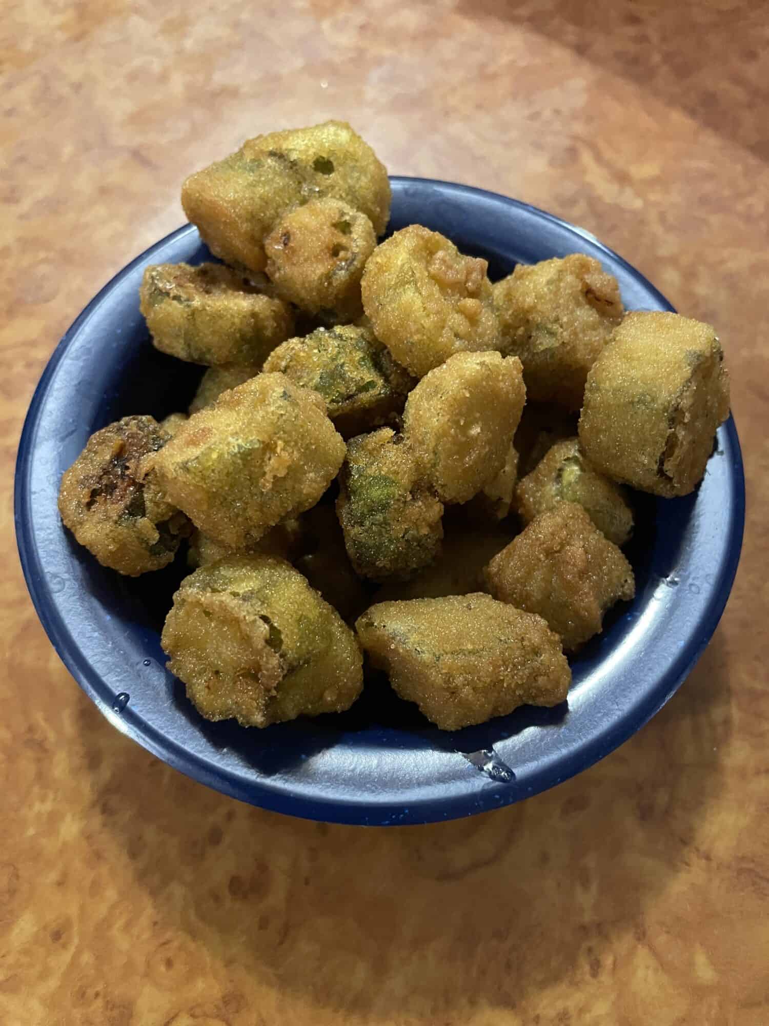 A small blue bowl of fried okra, cooked to a perfect golden brown with a brown table top background.