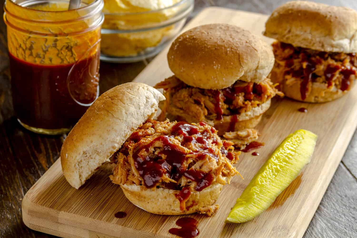 Three pulled pork barbeque sliders sitting on wooden cutting board with dill pickle with bottle of sauce and bowl of potato chips