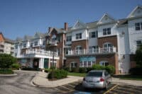 A large, multi-story building featuring brick on the lower floors and white horizontal siding on the upper floors, set against a clear blue sky. The building has multiple windows, several gabled roofs, and white balconies with intricate railings. In the foreground, a paved drive leads to a main entrance with white columns, and a parking lot with a silver sedan is visible. Green landscaping and bushes surround the property.