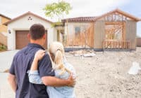 A couple, seen from behind, with the man's arm around the woman, stands on a dirt lot. They are looking towards two houses: one on the left is a completed, light-colored single-story home with a brown garage door and terracotta roof tiles, and the other on the right is a house under construction, framed with wood studs and partially clad in oriented strand board (OSB). A young tree stands between the two structures. The ground is rough dirt with some scattered construction debris.