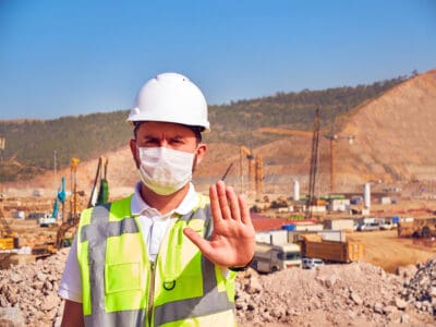 A male construction worker, facing forward, wears a white hard hat, a white disposable face mask, and a bright yellow and gray high-visibility vest. His right hand is raised, palm out, in a clear 'stop' gesture. Behind him, a large, dusty construction site is visible with piles of earth, heavy machinery including cranes and excavators, and distant hills under a clear blue sky.