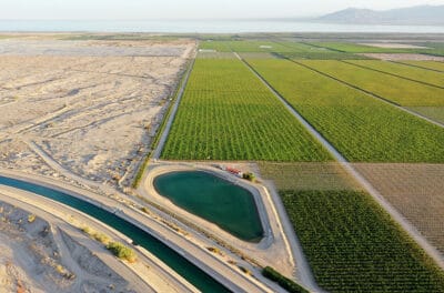 An aerial view captures a landscape sharply divided between lush green agricultural fields on the right and barren, sandy, arid land on the left. A winding irrigation canal with turquoise water runs diagonally across the lower-left portion, connecting to an irregularly shaped, bright teal reservoir. In the upper background, a large body of water, possibly the Salton Sea, stretches towards distant hazy mountains under a clear sky.