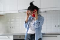A young Asian woman with dark hair tied in a messy bun stands in a modern, brightly lit white kitchen. She is looking down with a distressed expression, holding a red smartphone to her left ear with her right hand, while her left hand is pressed against her forehead. She wears a light blue open button-down shirt over a white t-shirt and dark jeans. The background shows white subway tiles, white kitchen cabinets, and a kitchen range hood.
