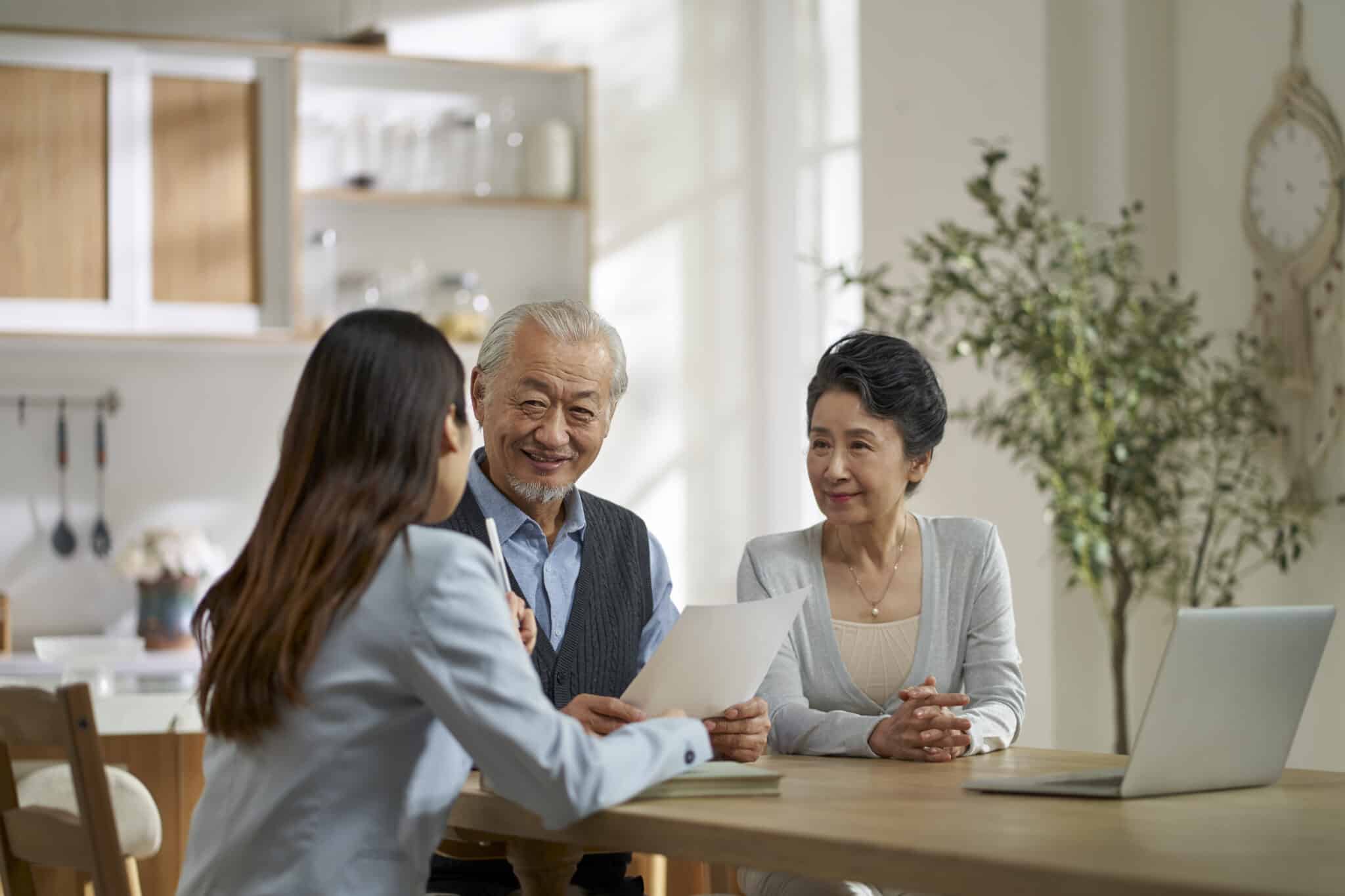 A young Asian woman with long dark hair, wearing a light blue blazer, sits at a wooden table across from an older Asian couple. She holds a pen and looks toward