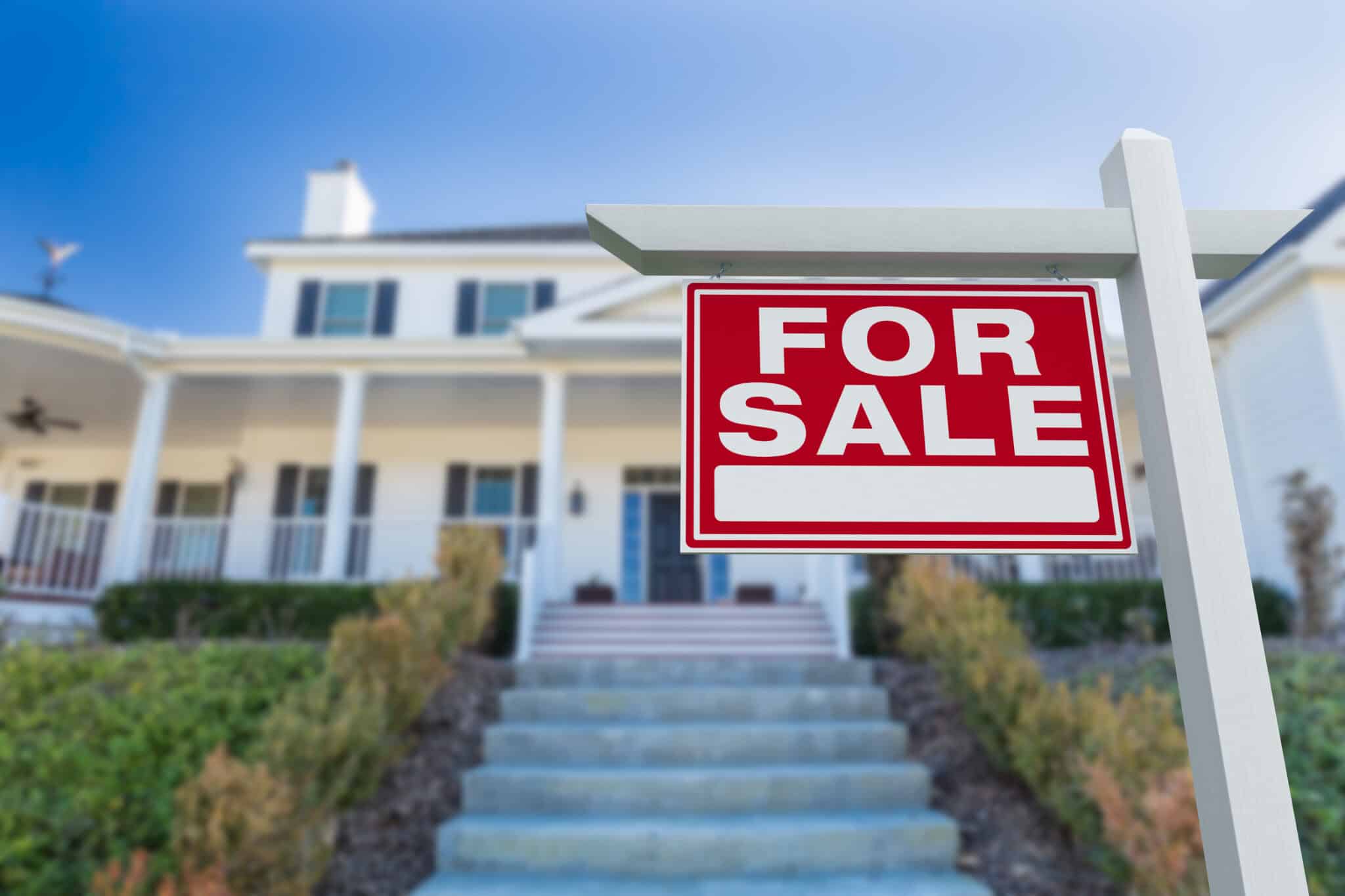 A red 'FOR SALE' sign with white lettering stands on a white post in the foreground. In the background, a large, blurred white house with dark shutters and a fr