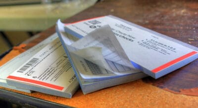 A close-up shot shows multiple stacks of blank white checkbooks resting on a textured brown wooden surface. The top checkbook in the foreground is slightly open, revealing a reorder form with text like 'Order Checks Now,' 'NOT NEGOTIABLE,' and instructions 'It's as easy as 1-2-3.' Red and blue stripes are visible along the edges of some checkbooks. Barcodes and partial account numbers are also discernible on some of the forms.
