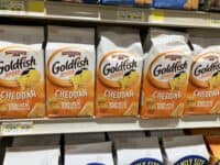 A close-up, eye-level photograph shows multiple white and orange bags of Pepperidge Farm Goldfish Cheddar Baked Snack Crackers arranged on a grocery store shelf. Each bag prominently features the Goldfish logo with a yellow fish wearing black sunglasses, along with text indicating 'Baked Snack Crackers', 'Cheddar', '100% REAL CHEESE', and 'NET WT 6.6 OZ (187g)'. Above the products, several yellow and white price tags are visible, displaying product details like 'Pepperidge Farm Goldfish Cheddar', various SKU numbers, a price of '$3.69', and several 'SELL BY' dates from 2024. The background features more retail shelving and products, slightly blurred.
