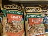 Two large, clear plastic bags of Snyder's of Hanover 'Family Size! Snaps Pretzels' are displayed on a silver wire shelf in a retail setting. The pretzel bags feature a dark brown and gold top section with 'Snyder's of Hanover' branding, transitioning to a bright green band with 'FAMILY SIZE!' and an orange lower section stating 'Snaps Pretzels'. The bags are filled with small, grid-shaped pretzels, clearly visible through the plastic. A third, partially visible bag of 'Bit-Size Pretzels' is to the right.