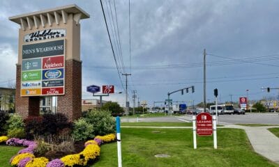 A tall brick and beige sign for 'Walden Crossings' shopping center, listing tenants like Men's Wearhouse, Starbucks, Applebee's, and The Vitamin Shoppe, stands amidst green grass and beds of yellow and purple flowers. In the background, an overcast sky hangs over a busy street intersection with traffic lights, utility poles, and signs for other businesses like Target, McDonald's, and Hampton Inn. A red and white 'LEASING' sign is visible in the foreground.