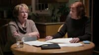 Two older women sit at a wooden kitchen table. The woman on the left, with blonde hair and glasses, wears a tan cowl-neck sweater and has her arms crossed, looking intently at the woman on the right. The woman on the right, with reddish-brown hair, wears a dark ribbed sweater, holds a pen, and looks down at the papers spread across the table. Documents, a black notebook, a glass of water, and a light blue smartphone are visible on the table. A kitchen with cabinets and a window is in the softly lit background.