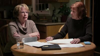 Two older women sit at a wooden kitchen table. The woman on the left, with blonde hair and glasses, wears a tan cowl-neck sweater and has her arms crossed, looking intently at the woman on the right. The woman on the right, with reddish-brown hair, wears a dark ribbed sweater, holds a pen, and looks down at the papers spread across the table. Documents, a black notebook, a glass of water, and a light blue smartphone are visible on the table. A kitchen with cabinets and a window is in the softly lit background.