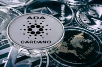 A close-up shot of several metallic cryptocurrency coins. A silver Cardano (ADA) coin, featuring its black dots logo and the text 'ADA CARDANO,' is in sharp focus at the center. Around it, other coins are partially visible and blurred, including a dark coin with 'ethereum' written vertically, another dark coin with a textured world map and 'ripple' text, and a silver Litecoin coin with a stylized 'L' symbol. The overall image has a cool, metallic tone.