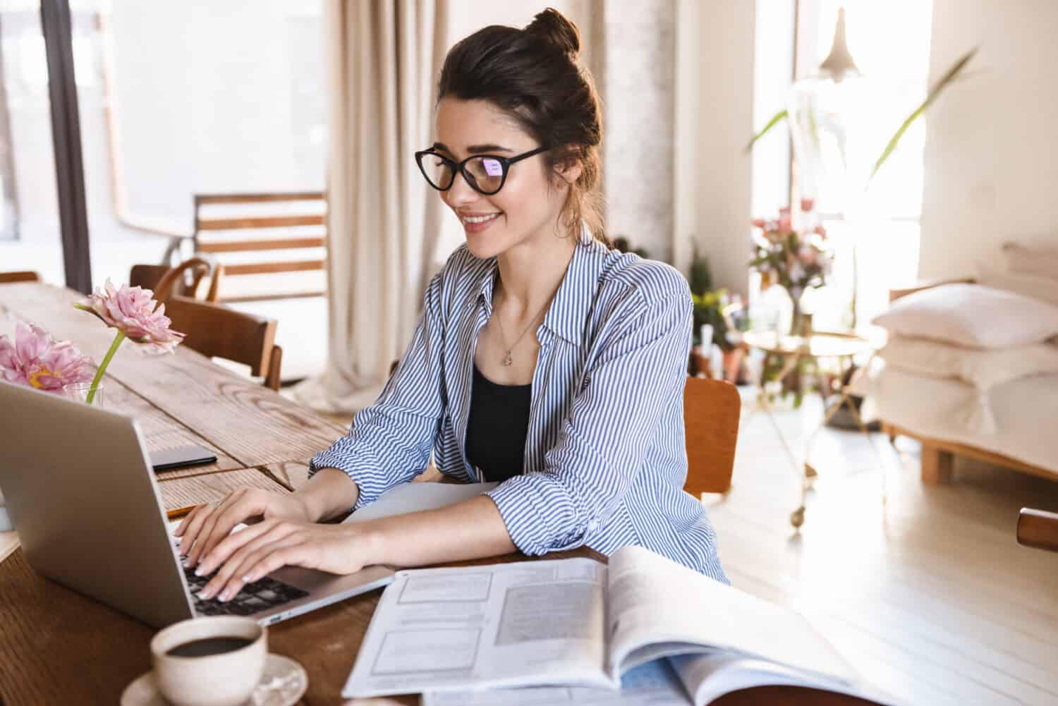 Image of smart positive woman 20s in casual clothing typing on laptop while working or studying at home