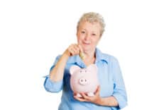 Closeup portrait of smiling senior mature woman depositing money into piggy bank, isolated on white background. Smart currency financial investment wealth decisions. Budget management and savings