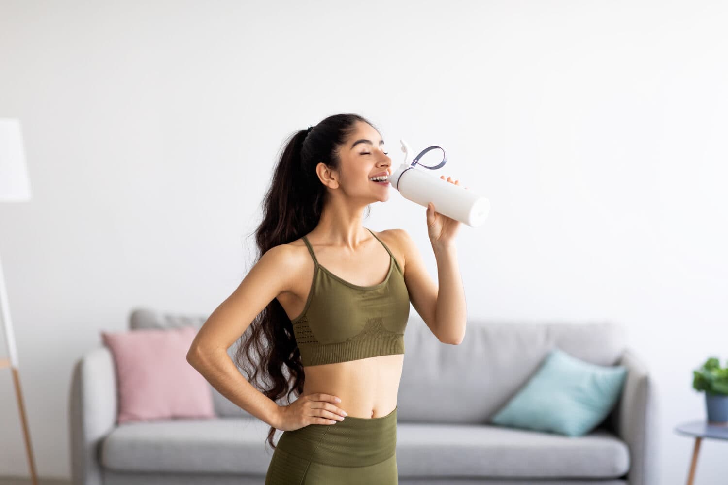 Pretty Indian woman in sports clothes drinking water or protein shake from bottle at home. Millennial Asian lady losing weight, keeping hydrated after workout, taking care of her body indoors