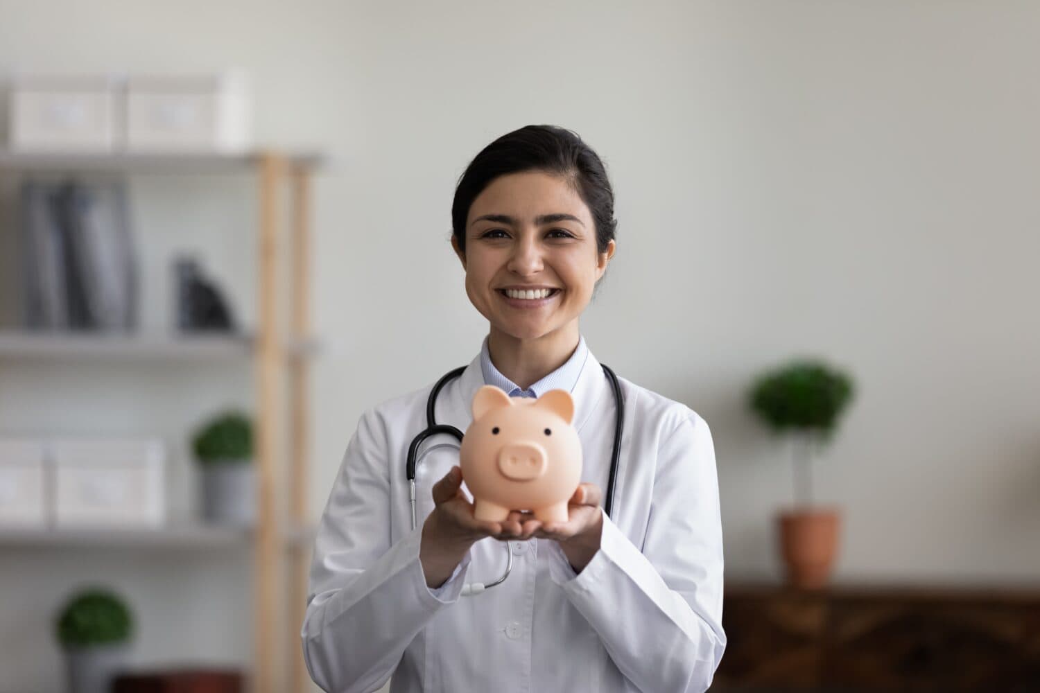 Head shot portrait smiling Indian female doctor physician in white uniform with stethoscope holding pink piggy bank, looking at camera, medical insurance or charity concept, healthcare and medicine