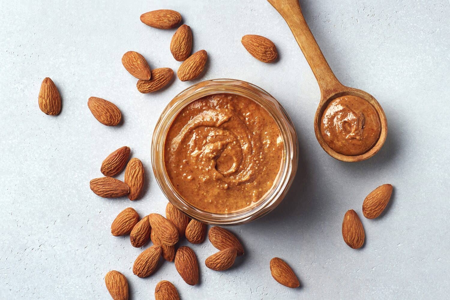 Glass jar and spoon of almond butter on light gray background, top view, above