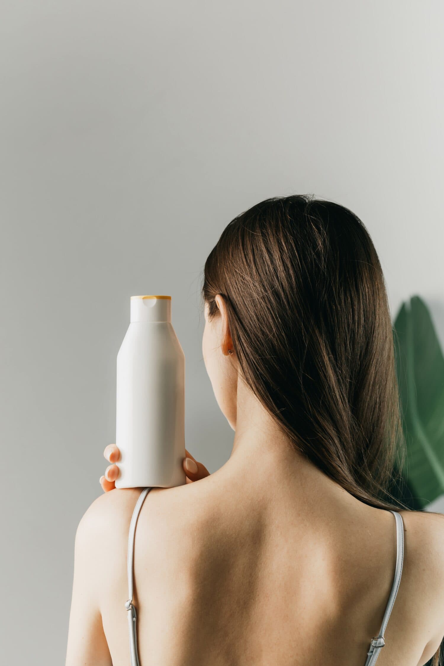 Woman holds a white bottle of cosmetics in her hand, standing with her back to the camera. Mock up shampoo, gel.