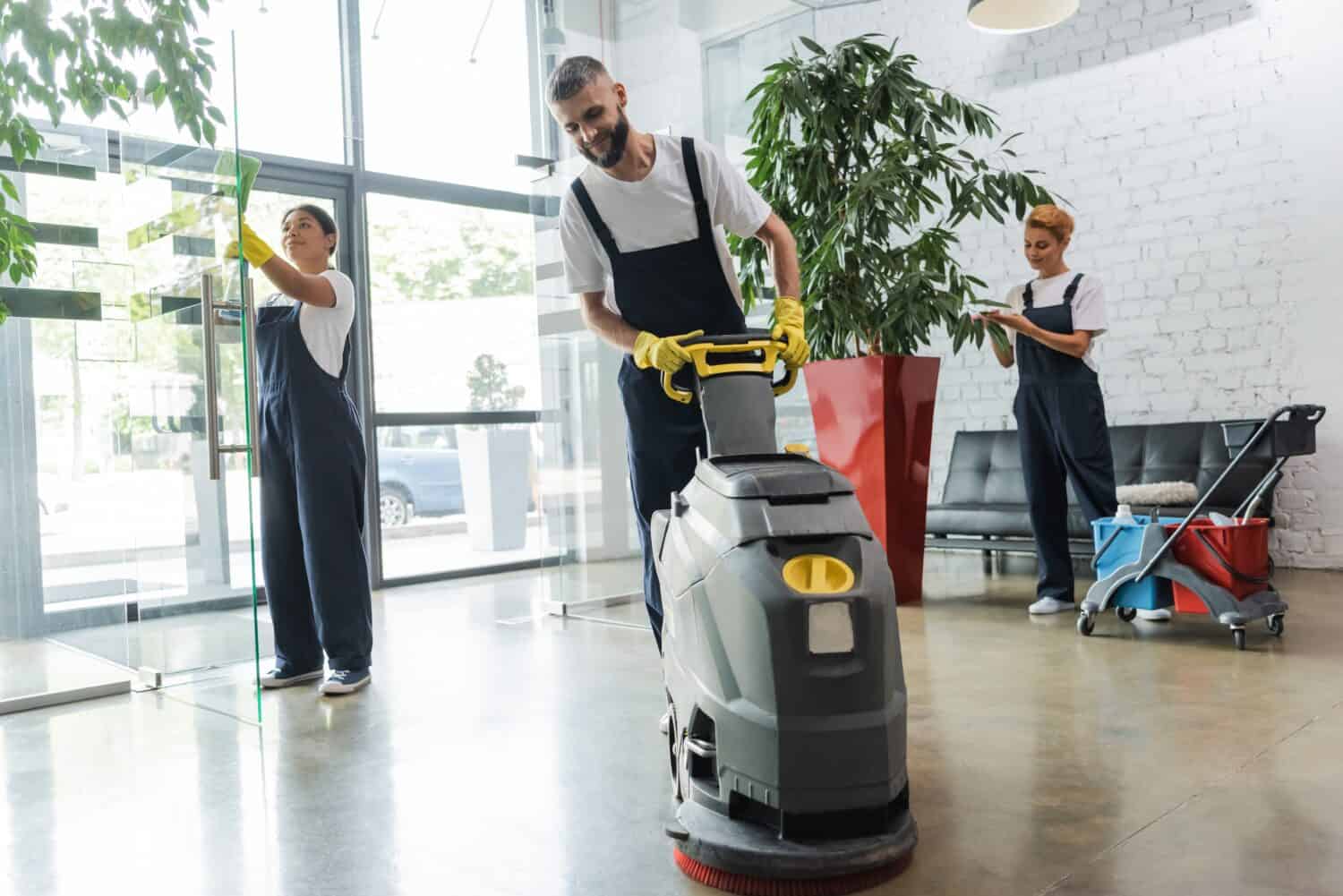 man in workwear with floor scrubber machine near interracial women cleaning office lobby