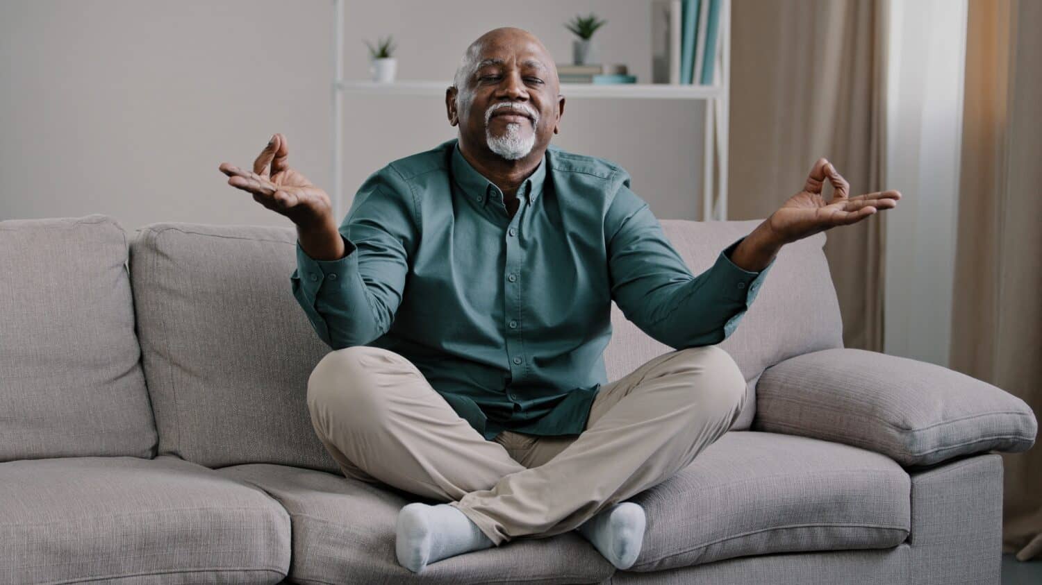 Happy elderly african american man sit on sofa in lotus position with folded fingers grandpa is meditating finds harmony relieves stress positive calm pensioner relaxing at home improve self-conscious