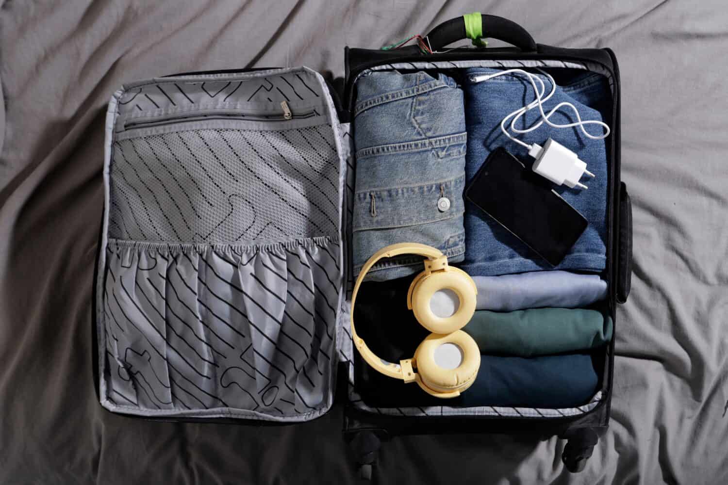 carry-on suitcase with blue clothes and large headphones, charger and cell phone on the bed