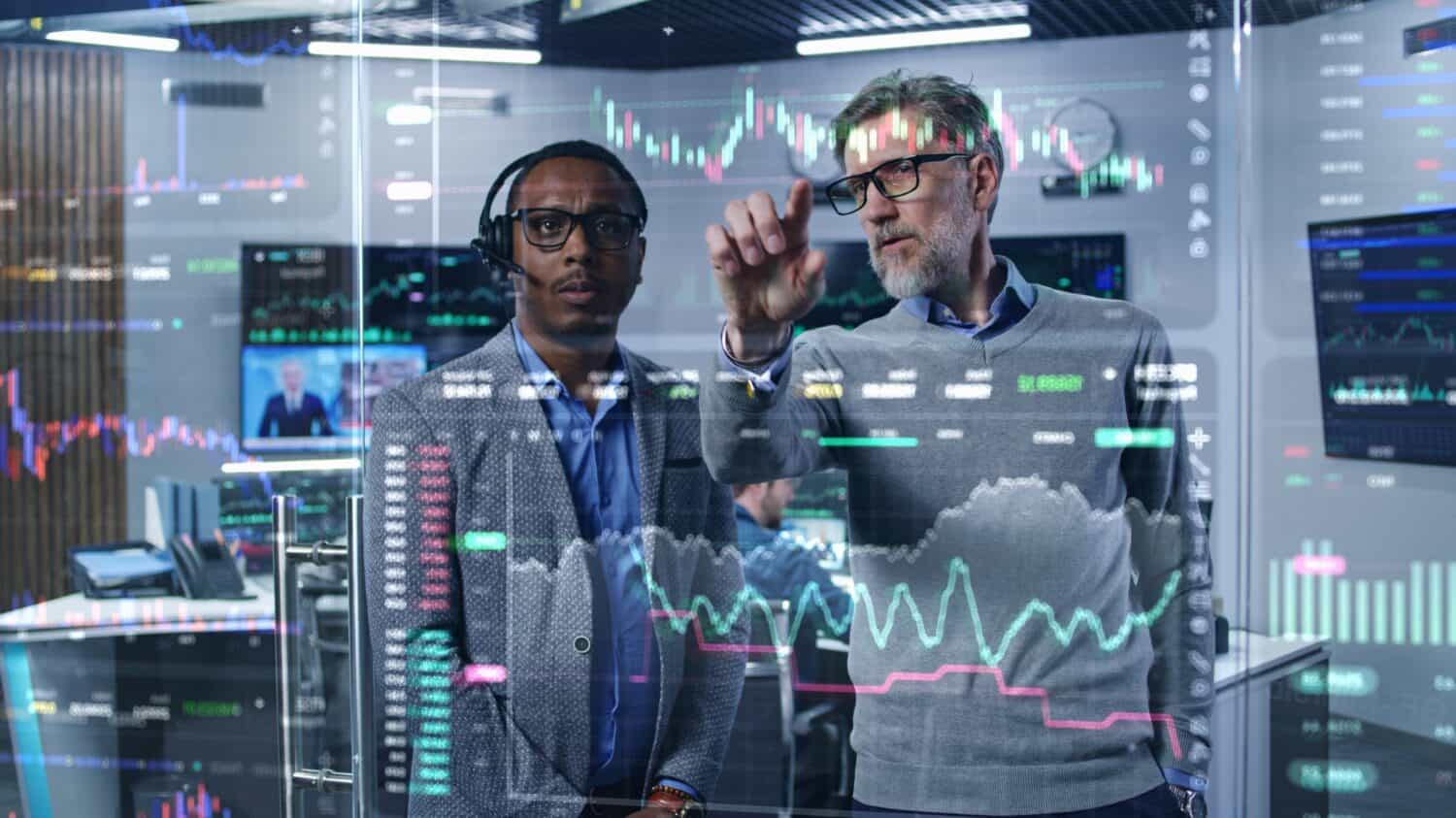 Multiracial financial analysts look on computer generated virtual 3D real-time stocks on glass wall, analyze business strategy in investment bank. Computers and big digital screens on background.