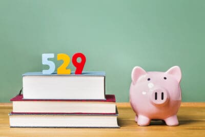 A light blue '5', a yellow '2', and a red '9' sit on top of a stack of three books. To the right, a pink ceramic piggy bank with black eyes and nostrils is on a wooden surface against a light green wall.