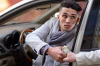 A young man with dark hair, wearing a light gray jacket and scarf, is seated in a car, extending his right hand out the window to receive a wad of money from another person's hand. He looks cautiously upwards and to the side, suggesting a secretive or high-stakes transaction. The interior of the car, including part of the steering wheel, is visible in the background.