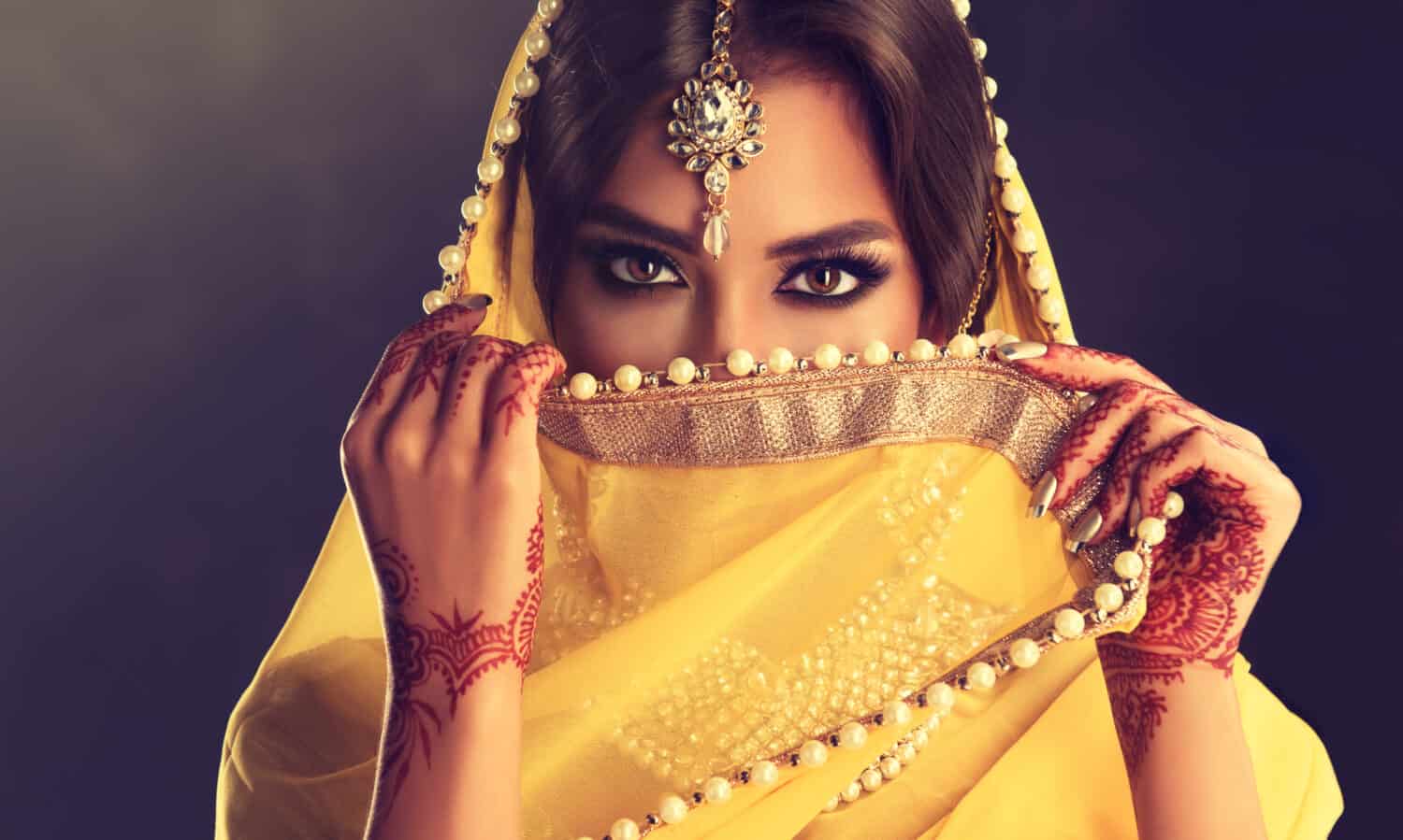 Beautiful indian girl . Young hindu woman model with tatoo mehndi and kundan jewelry . Traditional Indian costume yellow saree . Indian or Muslim woman covers her face.