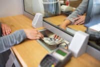A close-up view of a bank transaction, showing a customer's hands in a grey sleeve pushing a stack of hundred-dollar bills across a metallic tray towards a bank teller's hands, visible behind a clear protective screen. The counter is made of light wood, with a blurred payment terminal in the foreground.