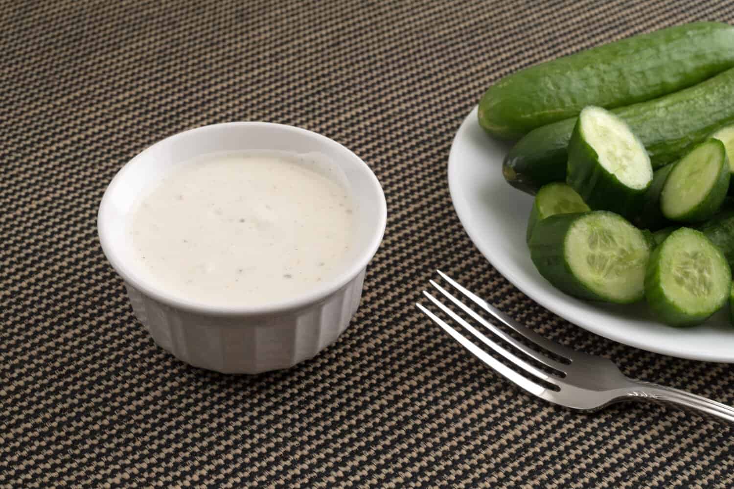 A small bowl of ranch dressing with sliced and whole bite size cucumbers on a white plate plus a fork in the foreground on a tablecloth.