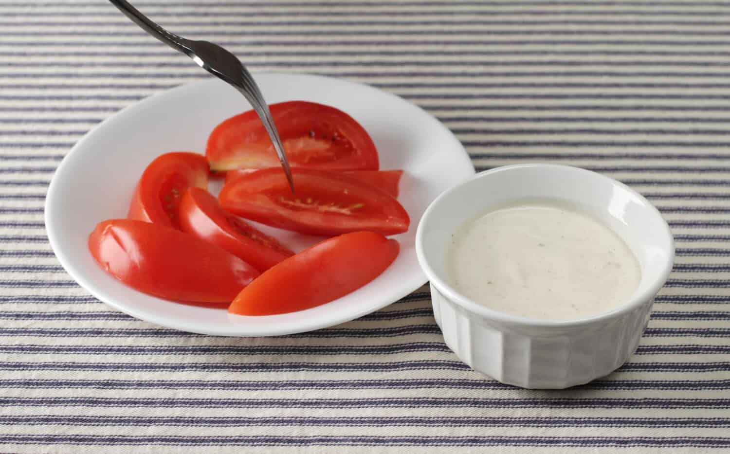 A white plate with slices of roma tomatoes with one slice being skewered with fork plus a bowl of ranch dressing to the side.