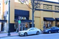 Street view of a vibrant yellow building housing a CAVA restaurant, with 'CAVA' in large white letters above the entrance at address 8515. Dark blue awnings and window frames contrast the yellow facade. A silver Mercedes-Benz and a blue Honda Civic are parked at the curb in front. A bare tree stands in front, and a person walks along the sidewalk to the right. A black street lamp with an ornate base is visible on the left.